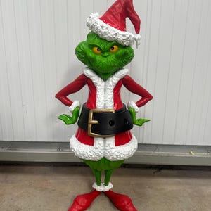 May include: A green figure of a character wearing a red Santa hat with white trim, a red coat with white trim, and a black belt with a gold buckle. The figure has green hands and feet, and is standing in a festive pose.