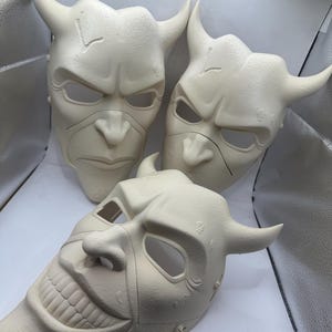 May include: Three off-white, horned masks with menacing expressions. The masks have eye and mouth openings, and the horns curve upwards. One mask has a wide, toothy grin. The masks appear to be made of a solid material.