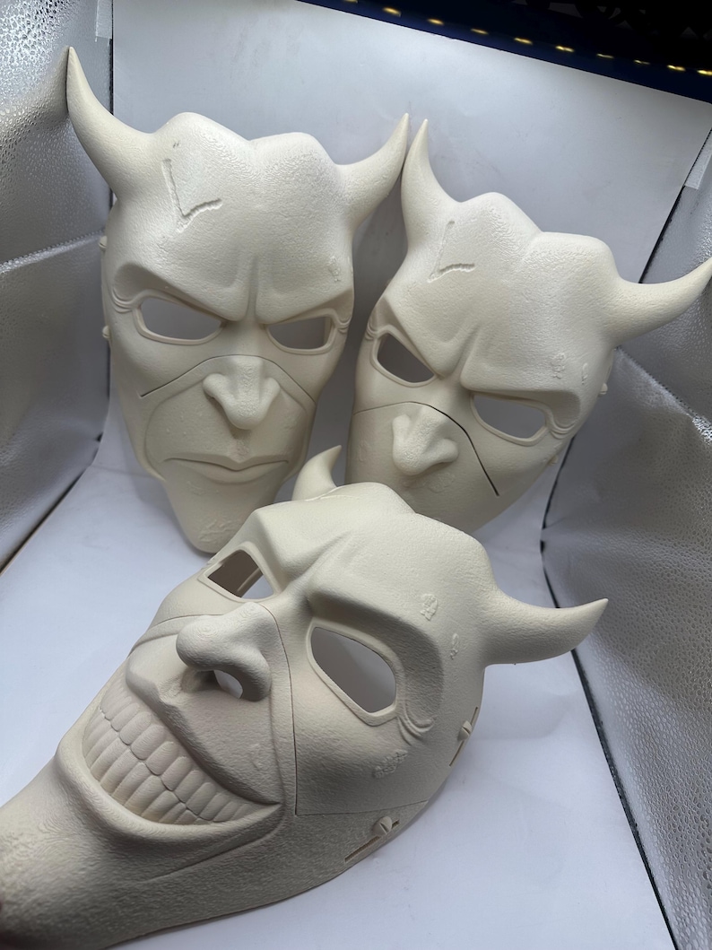May include: Three off-white, horned masks with menacing expressions. The masks have eye and mouth openings, and the horns curve upwards. One mask has a wide, toothy grin. The masks appear to be made of a solid material.