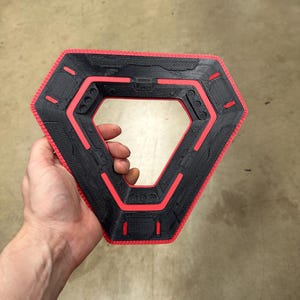 May include: A black and red geometric object with a hexagonal outer shape and a triangular inner opening. The object has a textured surface and red accents. The item is held in a person's hand.