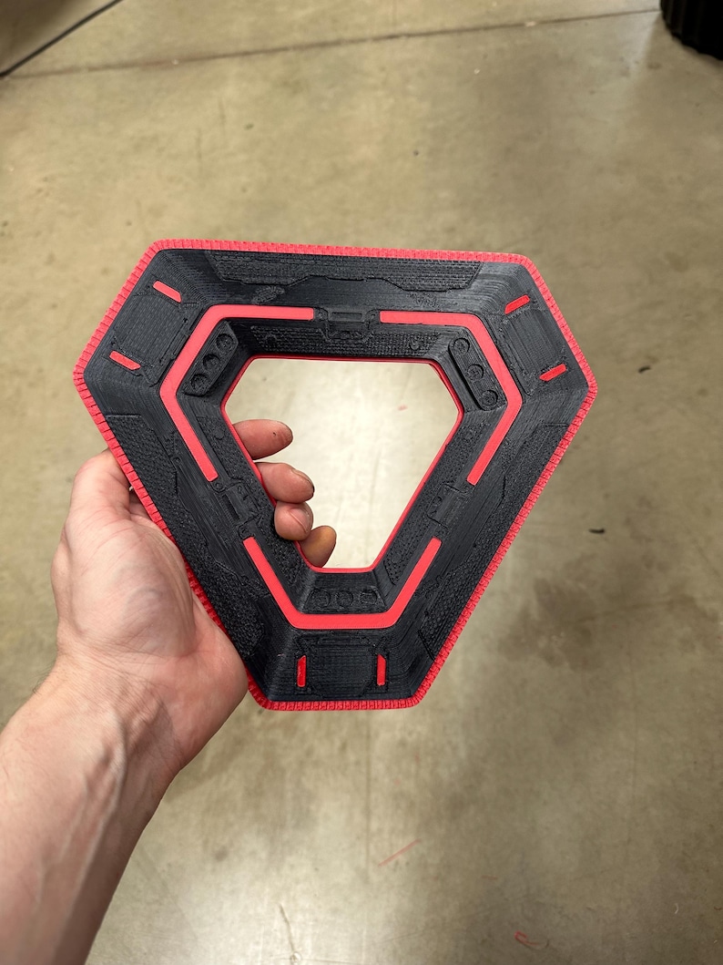 May include: A black and red geometric object with a hexagonal outer shape and a triangular inner opening. The object has a textured surface and red accents. The item is held in a person's hand.