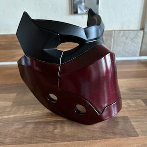 May include: A burgundy and black plastic mask with a geometric design. The mask has two holes for the eyes and two smaller holes on the lower part of the mask.