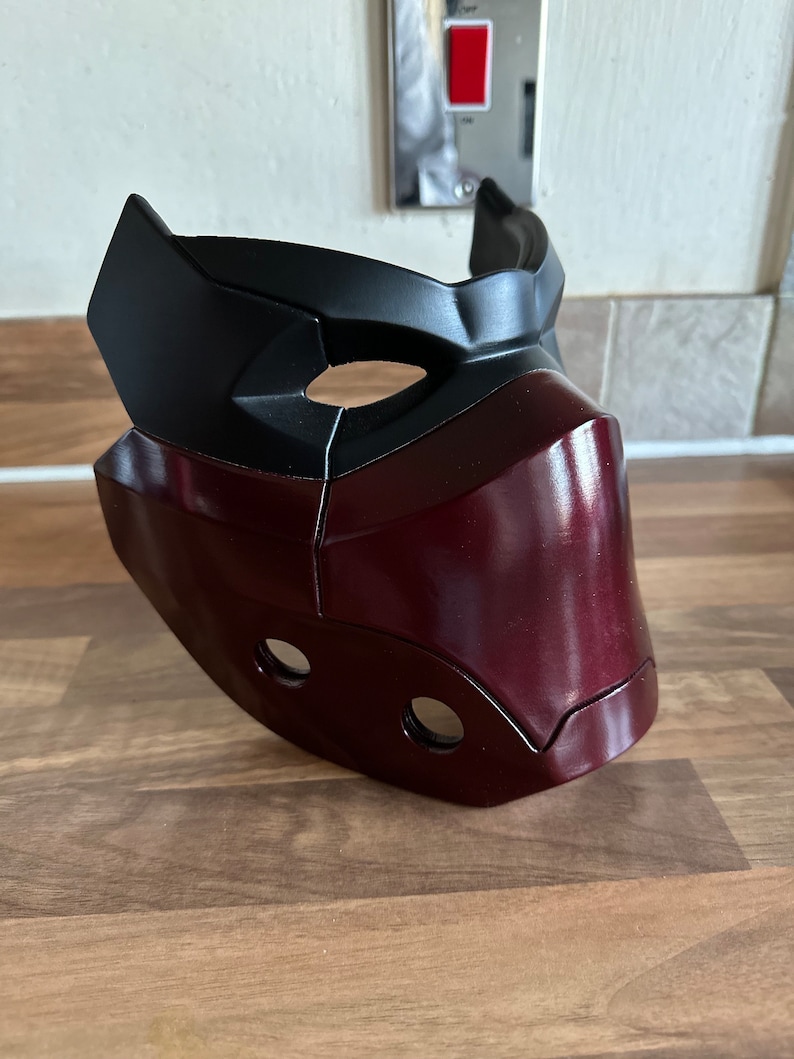 May include: A burgundy and black plastic mask with a geometric design. The mask has two holes for the eyes and two smaller holes on the lower part of the mask.