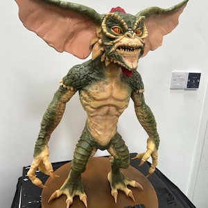 May include: A detailed sculpture of a Gremlin creature, featuring a green and tan color scheme with large, pink-toned ears. The figure has sharp teeth, red eyes, and a red crest. The base is brown with the word "GREMLINS" in black.