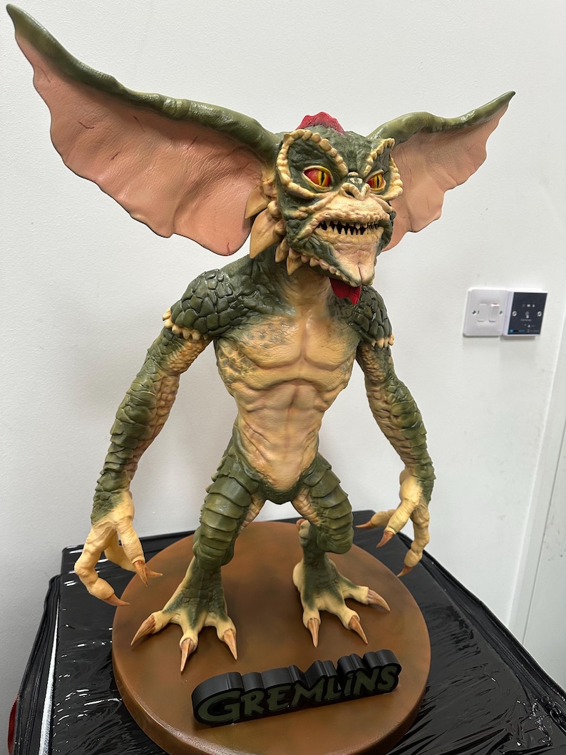 May include: A detailed sculpture of a Gremlin creature, featuring a green and tan color scheme with large, pink-toned ears. The figure has sharp teeth, red eyes, and a red crest. The base is brown with the word "GREMLINS" in black.