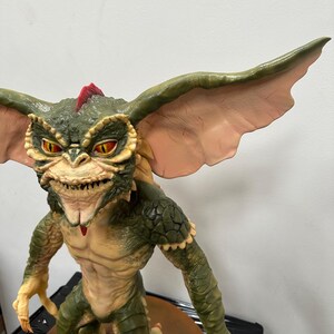 May include: A detailed figurine of a Gremlin, featuring a green and tan color scheme. The creature has large ears, sharp teeth, and red eyes. The figure is posed in a standing position, showcasing its claws and textured skin. The overall design captures the iconic look of the character.