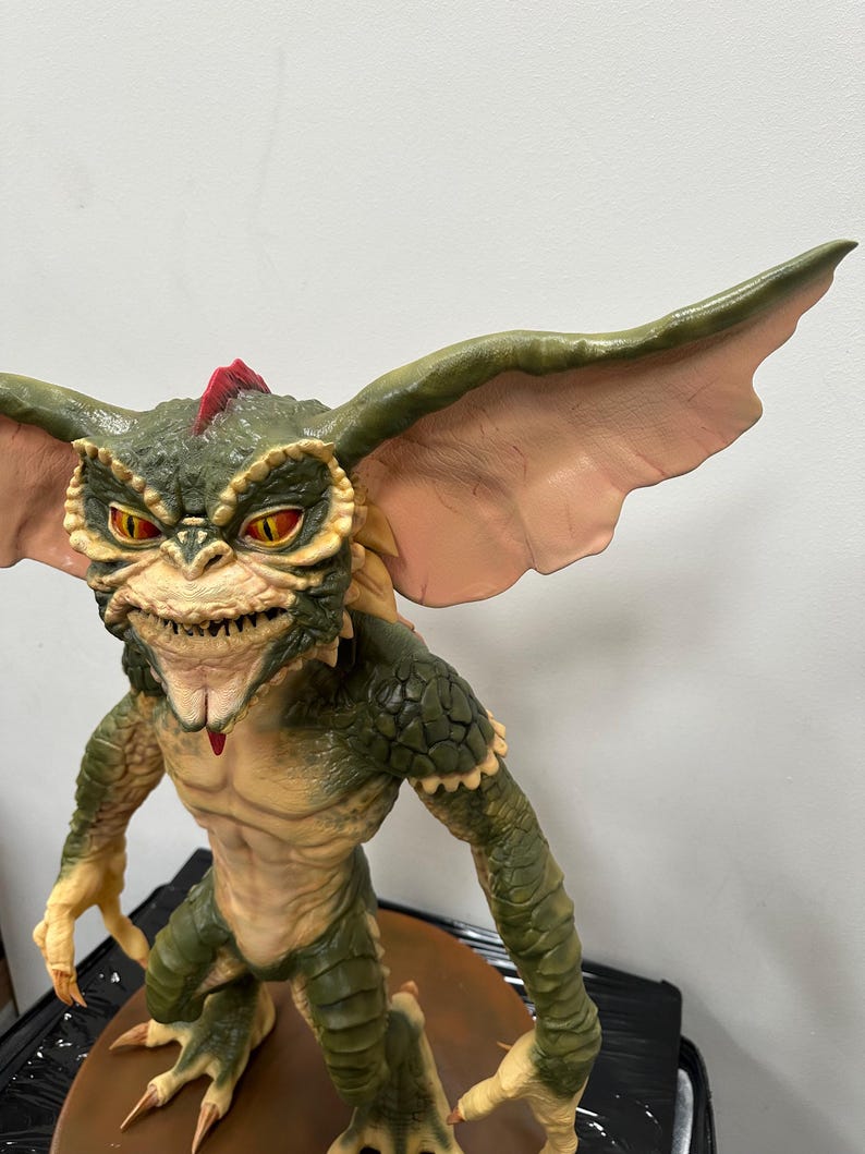 May include: A detailed figurine of a Gremlin, featuring a green and tan color scheme. The creature has large ears, sharp teeth, and red eyes. The figure is posed in a standing position, showcasing its claws and textured skin. The overall design captures the iconic look of the character.