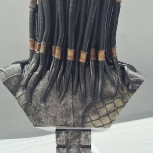 Puede incluir: Busto detallado de un alien&iacute;gena Predator, con rastas negras y detalles en marr&oacute;n. La escultura tiene un aspecto met&aacute;lico envejecido con intrincados detalles de armadura. Montado sobre una base geom&eacute;trica texturizada.