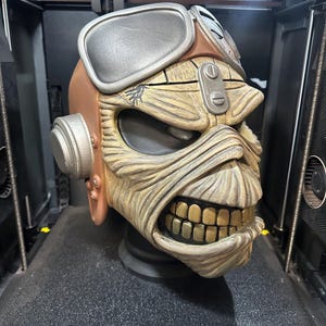 May include: A detailed, 3D-printed sculpture of a skull with aviator goggles and a brown helmet. The skull has a weathered appearance with visible teeth and is set against a dark background. The sculpture is likely a collectible or decorative item.