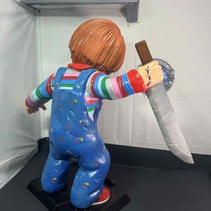 May include: A Chucky doll figurine, featuring a red-haired character in blue overalls with colorful striped sleeves, holding a large knife. The figurine stands on a black base with red accents, showcasing a detailed and menacing design.