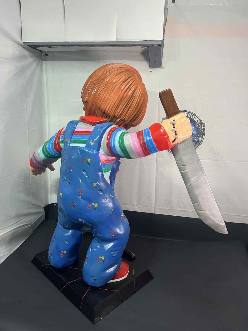 May include: A Chucky doll figurine, featuring a red-haired character in blue overalls with colorful striped sleeves, holding a large knife. The figurine stands on a black base with red accents, showcasing a detailed and menacing design.