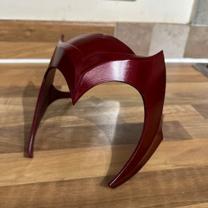 May include: A burgundy red plastic headband with a curved design. The headband is made of a smooth, glossy material.