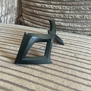 May include: A black plastic mask with a geometric design. The mask is lying on a brown striped fabric surface.