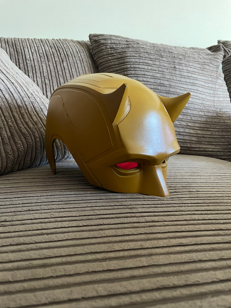 May include: A yellow Daredevil mask with red eyes. The mask is made of plastic and has a textured surface.