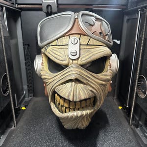 May include: A detailed, 3D-printed sculpture of a character's head, featuring a weathered appearance. The sculpture has a grinning mouth with visible teeth, goggles, and a respirator. The color palette includes shades of brown, tan, and gray.