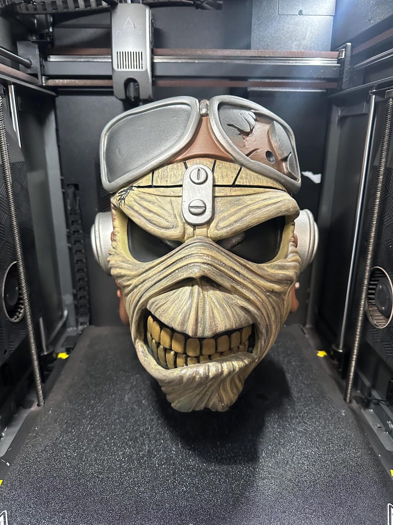May include: A detailed, 3D-printed sculpture of a character's head, featuring a weathered appearance. The sculpture has a grinning mouth with visible teeth, goggles, and a respirator. The color palette includes shades of brown, tan, and gray.