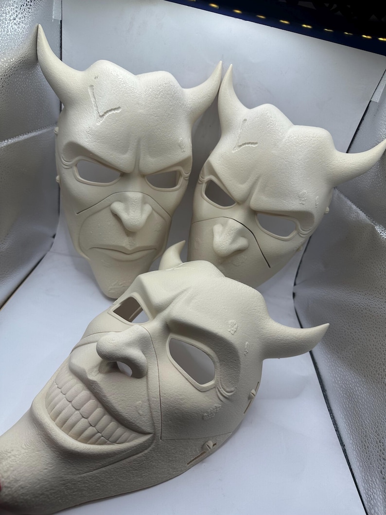 May include: Off-white, horned masks with menacing expressions. Three masks are displayed, each with a unique facial expression. The masks have eye and mouth openings. The masks have a textured surface and are likely made of a durable material.