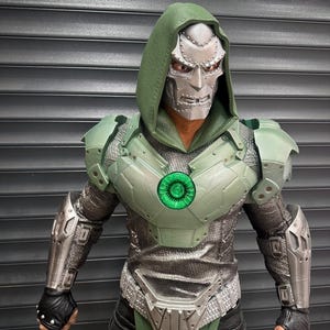 May include: A detailed action figure of a character in a green and silver armored suit with a silver mask and green hood. The figure has a glowing green chest emblem and black gloves. The background is a gray, slatted surface.