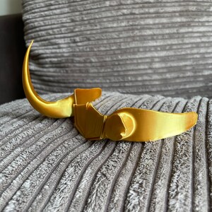 May include: A gold-colored plastic headband with a curved design, resembling a crown or a headdress. The headband is on a gray textured surface.