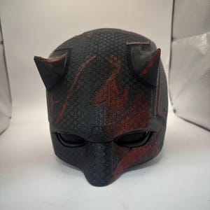 May include: A black and red helmet with a textured surface, featuring two pointed horns on top and eye openings. The helmet has a dark color scheme with red accents, resembling a superhero mask.