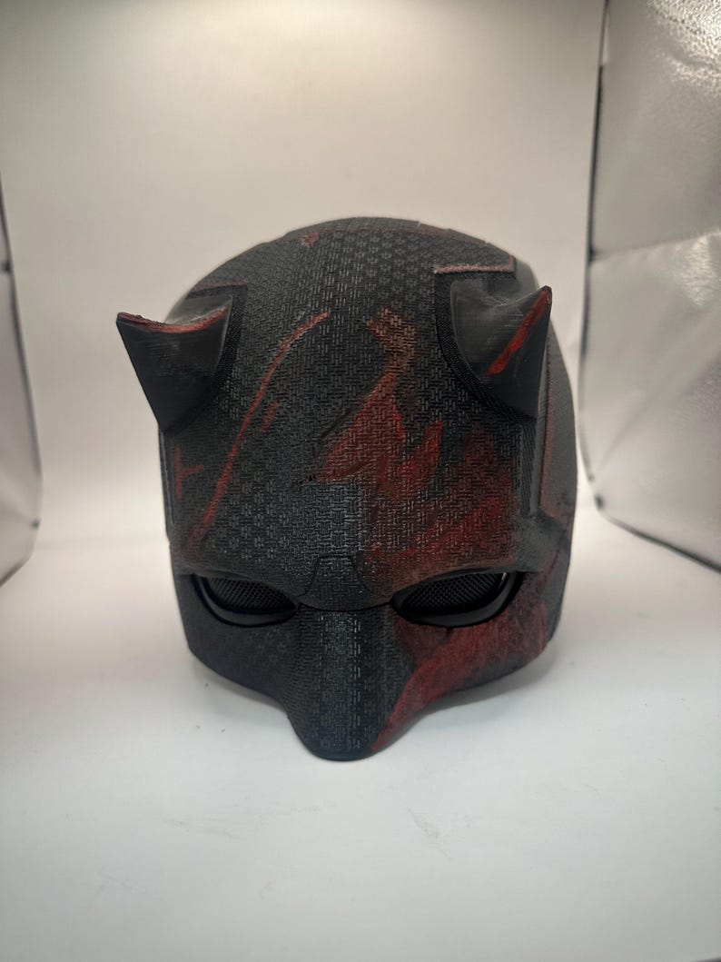 May include: A black and red helmet with a textured surface, featuring two pointed horns on top and eye openings. The helmet has a dark color scheme with red accents, resembling a superhero mask.