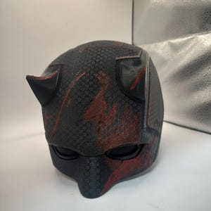 May include: A black and red helmet with a textured surface and two pointed horns. The helmet has mesh eye coverings and red paint splatters. The design is reminiscent of a superhero costume.