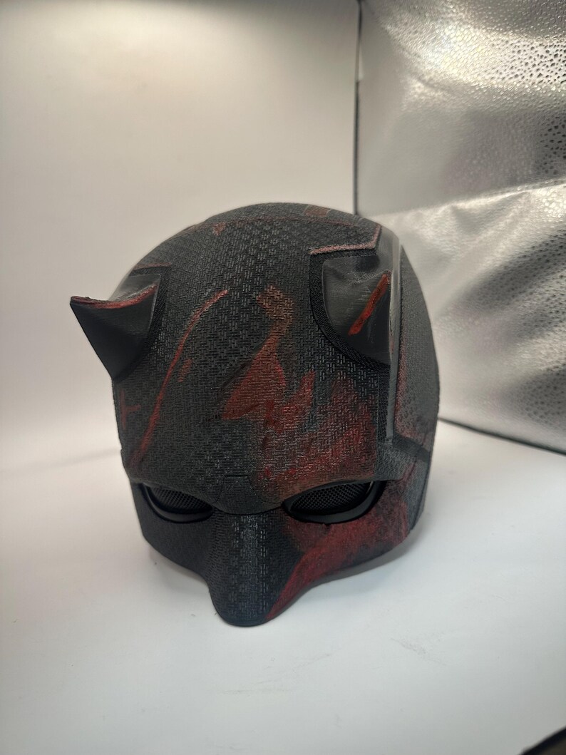May include: A black and red helmet with a textured surface and two pointed horns. The helmet has mesh eye coverings and red paint splatters. The design is reminiscent of a superhero costume.