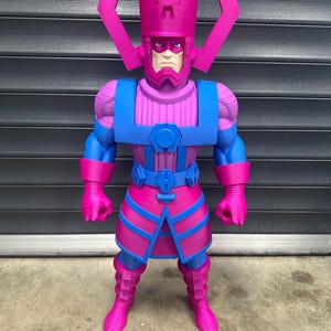 May include: A large, colorful action figure of Galactus, a comic book character. The figure is predominantly pink and blue, with a detailed helmet and armor. The figure stands on a concrete surface against a dark gray background.