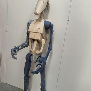 May include: A beige and blue articulated robot figure with a humanoid shape. The robot has a large head with a rounded top and a small, rectangular mouth. The robot's arms and legs are made of multiple segments, and the joints are visible. The robot is standing on a white surface.