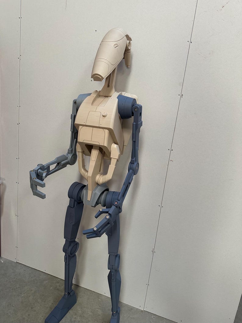 May include: A beige and blue articulated robot figure with a humanoid shape. The robot has a large head with a rounded top and a small, rectangular mouth. The robot's arms and legs are made of multiple segments, and the joints are visible. The robot is standing on a white surface.