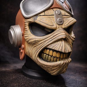 May include: A detailed sculpture of a skull with a menacing expression, featuring a weathered, textured appearance. The skull wears aviator goggles and a brown leather helmet. The teeth are a golden color, and the overall design is reminiscent of a rock and roll theme.