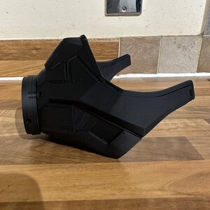 May include: A black plastic mask with a futuristic design. The mask has a large, round opening for the mouth and nose, and a smaller opening for the eyes. The mask is made of a smooth, matte plastic.