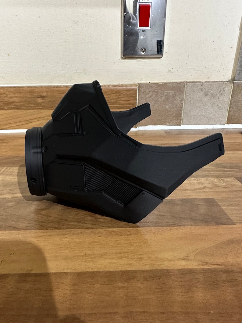 May include: A black plastic mask with a futuristic design. The mask has a large, round opening for the mouth and nose, and a smaller opening for the eyes. The mask is made of a smooth, matte plastic.