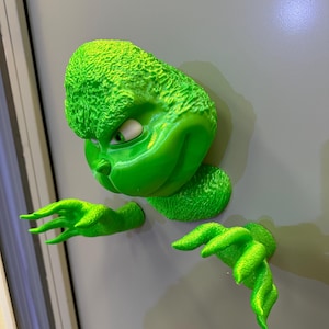 May include: A green 3D printed Grinch figure with a mischievous expression, peeking out from behind a door. The figure has a textured surface and is holding out its hands.