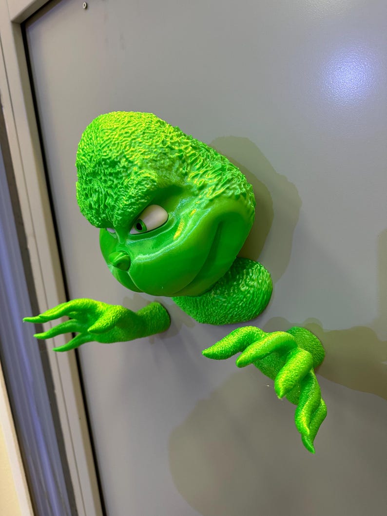 May include: A green 3D printed Grinch figure with a mischievous expression, peeking out from behind a door. The figure has a textured surface and is holding out its hands.