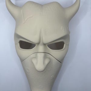 May include: A light beige mask with a devilish design. The mask features two curved horns, eye openings, and a prominent nose. The surface has a textured appearance with some imperfections. The mask is likely for costume or decorative purposes.