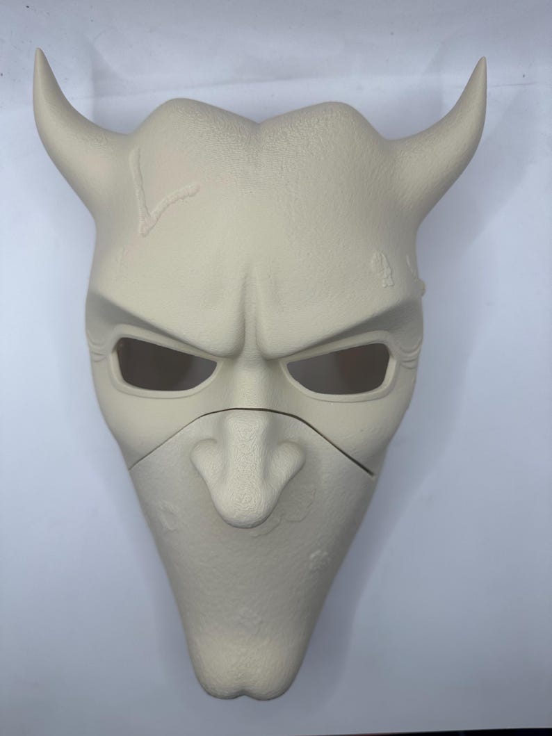 May include: A light beige mask with a devilish design. The mask features two curved horns, eye openings, and a prominent nose. The surface has a textured appearance with some imperfections. The mask is likely for costume or decorative purposes.