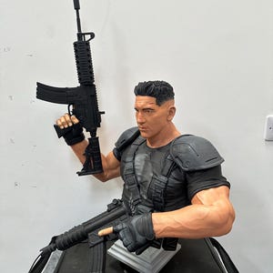 May include: A detailed bust of a character holding two black rifles. The figure is wearing a black tactical vest and gloves, with a skull emblem on the chest. The sculpture has a realistic, muscular physique and short, dark hair.