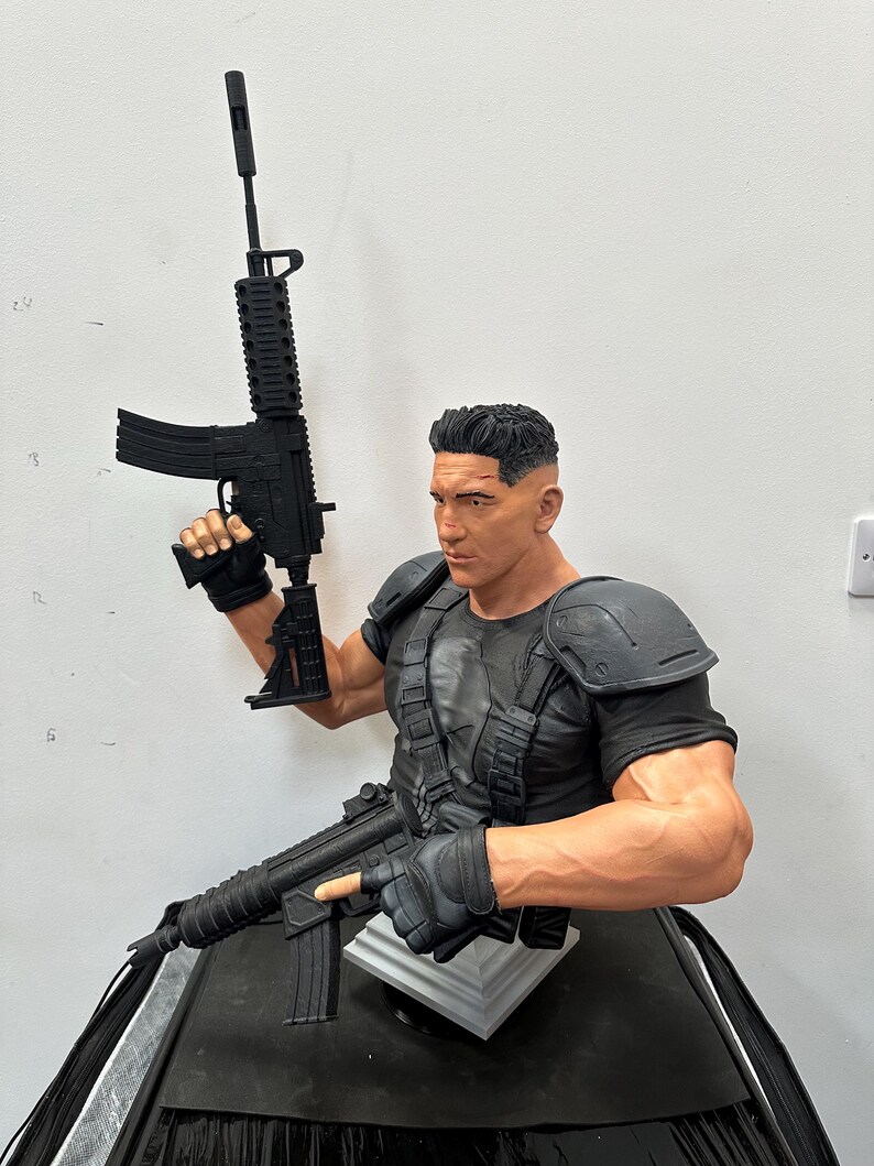 May include: A detailed bust of a character holding two black rifles. The figure is wearing a black tactical vest and gloves, with a skull emblem on the chest. The sculpture has a realistic, muscular physique and short, dark hair.