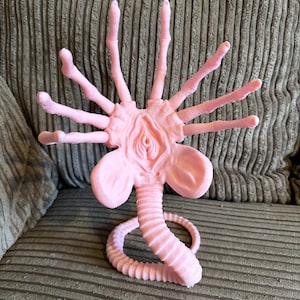 Op de afbeelding: Een roze plastic figuur van een buitenaards wezen met een lange, gesegmenteerde staart en meerdere armen. Het wezen heeft een groot, rond hoofd met een mond en twee ogen.
