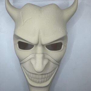 May include: A cream-colored mask with a sinister grin, curved horns, and eye openings. The mask has a textured surface and a crack on the forehead. The design is reminiscent of a demonic or villainous character.