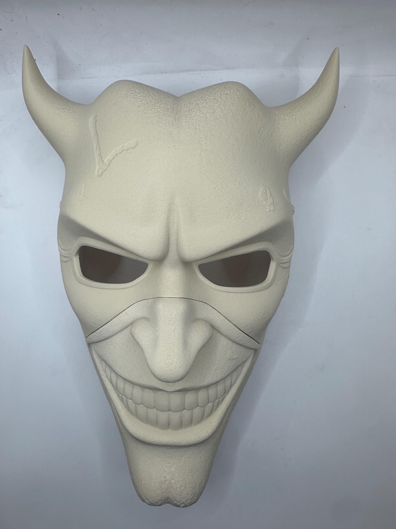 May include: A cream-colored mask with a sinister grin, curved horns, and eye openings. The mask has a textured surface and a crack on the forehead. The design is reminiscent of a demonic or villainous character.