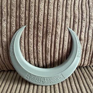 May include: A gray crescent moon-shaped object with intricate designs and text on both sides. The object is made of a smooth, plastic-like material.
