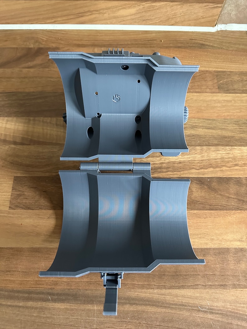 May include: A grey 3D printed object with a hinged lid and a small lever. The object is open and the inside is visible.