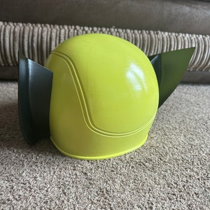 Pode incluir: Um capacete de pl&aacute;stico amarelo e preto com formato arredondado e duas pequenas orelhas. O capacete &eacute; projetado para se parecer com um personagem de desenho animado.