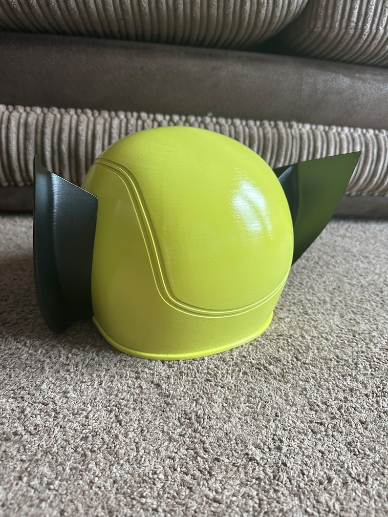 Pode incluir: Um capacete de pl&aacute;stico amarelo e preto com formato arredondado e duas pequenas orelhas. O capacete &eacute; projetado para se parecer com um personagem de desenho animado.