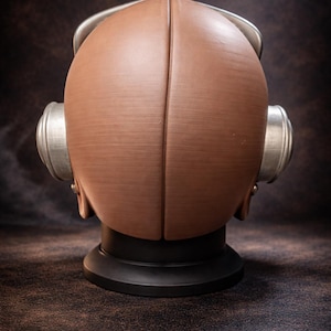 May include: A brown, retro-style diving helmet with silver accents. The helmet is made of two halves joined at the center, with circular ear pieces on each side. It sits on a black base, against a dark brown background.