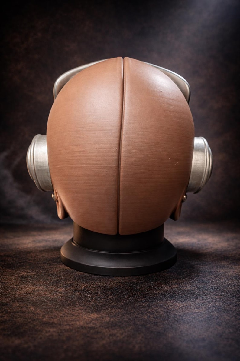 May include: A brown, retro-style diving helmet with silver accents. The helmet is made of two halves joined at the center, with circular ear pieces on each side. It sits on a black base, against a dark brown background.
