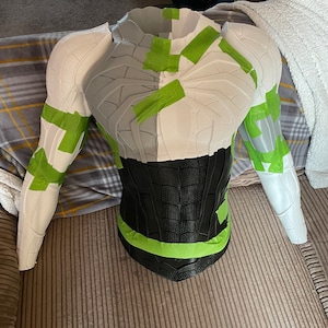 Puede incluir: Una pieza de armadura de torso texturizada, parcialmente pintada, con brazos adjuntos. La armadura es principalmente blanca y gris, con detalles en negro en la parte inferior. Cinta de pintor verde asegura las piezas. El art&iacute;culo est&aacute; sobre una superficie rayada.