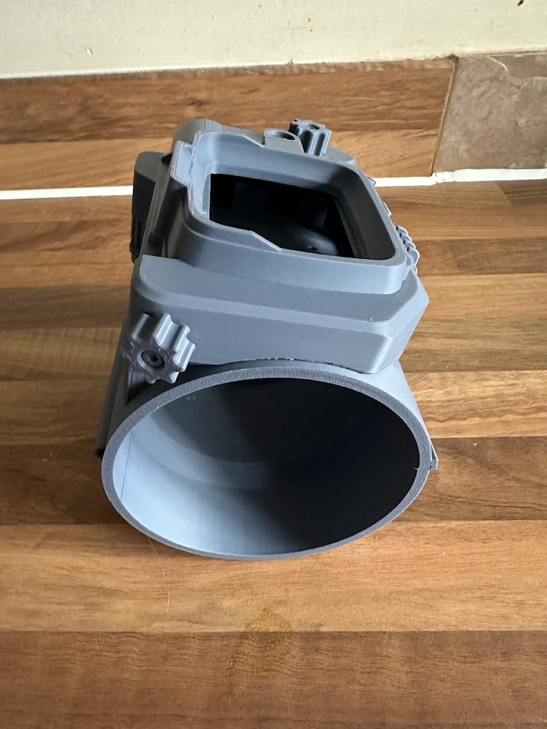 May include: A grey 3D printed camera cage with a round opening at the bottom and a rectangular opening at the top. The cage has two adjustable knobs on the side.